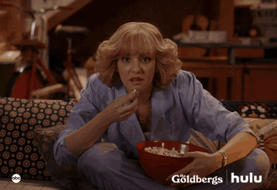 TV gif. Wendi McClendon Covey as Beverly in The Goldbergs, sits on a couch hunched over and eating out of a red bowl of popcorn. She leans forward with wide eyes glued to the screen ahead of her. 