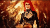 Music video gif. Britney Spears wears a black leather outfit, walking from a fire as her long red hair flies around her, in the video for her song "Toxic."
