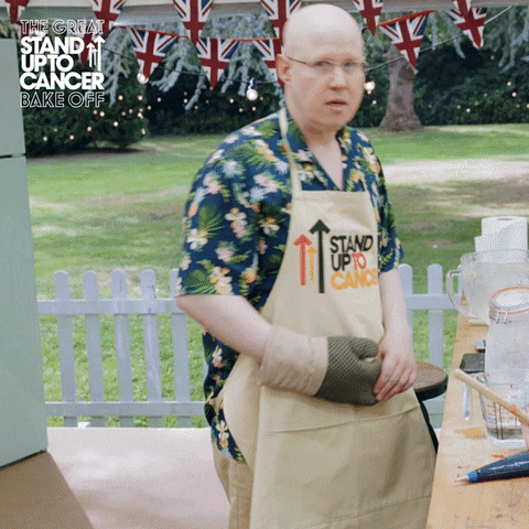 Reality TV gif. Matt Lucas from the Great British Bake-Off pulls off an oven mitt and raises two fingers in the air. He is wearing an apron that says "stand up to cancer."
