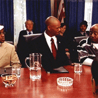 TV gif. Dave Chappelle as Black Bush at a conference desk shakes his head, then knocks over a pitcher of water and jumps up, causing chaos.