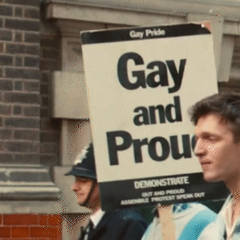 Movie gif. Young man in the movie "Pride" marches in a parade, holding a sign that reads, "Gay Pride: Gay and Proud. Demonstrate. Out and Proud. Assemble, protest, speak out." The scene flashes to an old woman holding a large sign in front of her chest that reads, "Burn in hell."