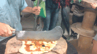 Video gif. Handmade pizza sits on a wooden log slab. A man holds a huge knife that almost looks like a sword or machete and then hits the pizza, instantly slicing it in half.
