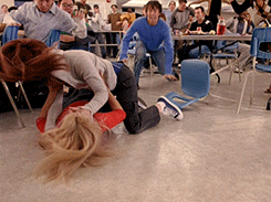 Movies gif. Cady and Regina in Mean Girls fight on the floor of the cafeteria. Cady is on top of Regina and they shake one another vigorously while the crowd cheers them on.
