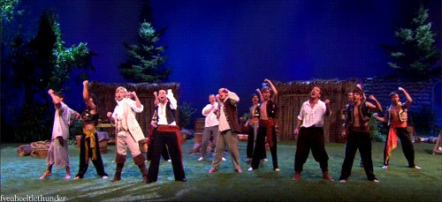 Video gif. On a stage set with large prop trees, a group of performers in period clothing raise their fists in unison. Text, "This."