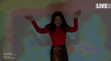 TV gif. Vivienne Tam on New York Fashion Week: The Show stands in front of a projection screen, video playing behind her, while she smiles and waves with her arms out to her sides, and her hands above her head. 