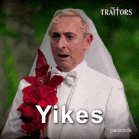 Sponsored gif. Alan Cumming as host of in The Traitors wearing a white suit with red flowers pinned to the left side and a white wedding veil grimaces and points. Text reads, “Yikes.”