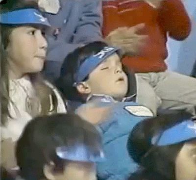 Video gif. A boy sits back in an audience of people who are cheering. He’s dead asleep with his mouth a little open. He sleeps through all the ruckus around him and even as the person sitting behind him leans over. 