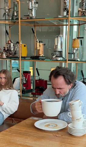 Video gif. A man in a coffee shop drinks from an enormous cup of coffee (we're talking huge, here) as a girl looks on in amazement. He's seated next to several empty coffee cups. That much coffee just wasn't enough for him, it seems.