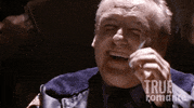 Movie gif. Dennis Hopper as Clifford in True Romance. He's beaten up and sitting on a chair, cackling. He raises his cigarette in toast to the person and takes a puff.