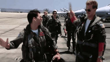 Movie gif. Tom Cruise as Maverick and Val Kilmer as Iceman in Top Gun walk along with a line of other pilots away from the planes. Maverick and Iceman high five excitedly. 