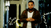 Video gif. A man glances down at his wrist watch and then taps his fingers together impatiently. 