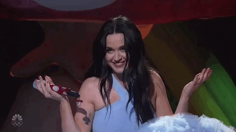 Celebrity gif. Katy Perry sits down on stage with a gown on. She holds a bedazzled microphone in one hand as she smiles at the crowd. She makes her empty hand into a peace sign and shakes it.