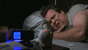 TV gif. Eric Stonestreet as Cam on Modern Family lies in bed in the dark looking at the clock on his bedside table, shaking as he cries. 