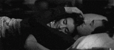 Video gif. Black and white footage of a couple cuddling in bed. The woman nestles into the man's shoulder as he runs his fingers through her hair.