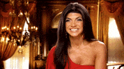 Reality TV gif. Teresa Giudice from Real Housewives of New Jersey. She waves eagerly while being interviewed and grins.