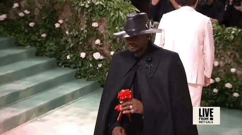 Met Gala 2024 gif. Usher, wearing an all-black custom Alexander McQueen ensemble, eyes spookily hidden by his fedora, holds a red paper flower.