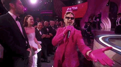 Oscars 2024 GIF. Ryan Gosling, dressed in a Barbie pink tuxedo and sunglasses, reaches out and grabs the disembodied hand of the camera man, holds it, kisses it, then leads him up onto the stage.