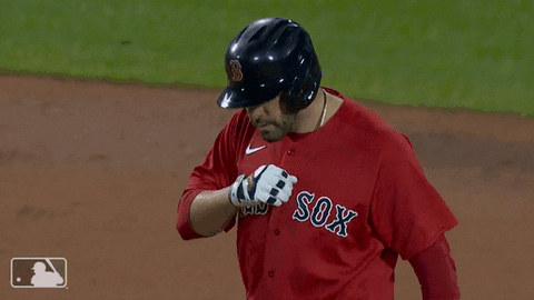 Sports gif. JD Martinez wears his Red Sox uniform on the field as he taps his chest, then looks up and points to the sky, before waving toward us.