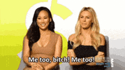 Video gif. Two women sit for an interview smiling, as one leans forward and, from the captions, enthusiastically says, "Me too, bitch! Me too!" The other girl laughs and holds her hand to her chest. 