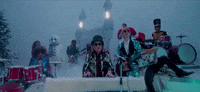 Music video gif. Elton John plays the piano and Ed Sheeran plays the guitar in the Merry Christmas music video. Snow falls around them and their band members. There’s a castle in the background.