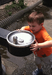 Video gif. A little boy pushes the button on a drinking fountain and closes his eyes as the water spurts out, spraying him in the face on and landing on his head.