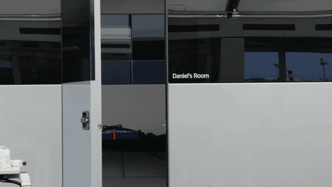 Sports gif. Daniel Ricciardo in his racing suit appears in a doorway, smiles and waves at us, then leaves to the right. Text printed next to the doorway marks the area as “Daniel’s Room.”