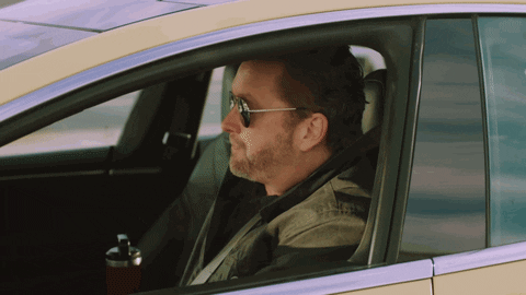 Video gif. A man wearing dark aviator sunglasses sits in the driver's seat of a car. Looking straight ahead, he grabs the wheel and the car zooms off. Text, "Have a great weekend everybody."
