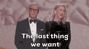 Oscars 2024 GIF. Catherine O'Hara and Michael Keaton stand at the podium, announcing the nominees for Best Makeup and Costume. O'Hara looks around the crowd and shrugs her shoulders as she sweetly says, "The last thing we want is anyone knowing what we really look like." Keaton solemnly nods in agreement. 