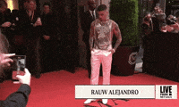 Met Gala 2024 gif. Rauw Alejandro wearing a golden sleeveless top and white satin pants, an ensemble by Ludovic de Saint Sernin, turns slowly, adjusting nervously, fully surrounded by cameras.