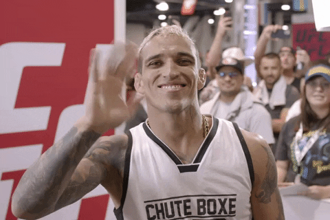 Sports gif. Charles Oliveira from UFC smiles cheekily at the camera while waving and squeezing his fingers cutely.