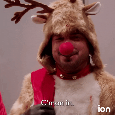 Video gif. A man dressed in a Rudolph the Red-Nosed Reindeer costume smiles and gestures toward himself as he says, "C'mon in."