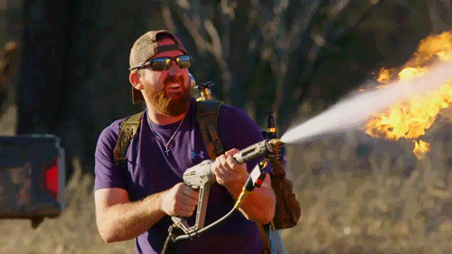 Reality TV gif. Bearded man on the Dude Perfect show wears a backwards baseball cap and sunglasses with a flamethrower on his back, spraying fire in front of him from side to side.