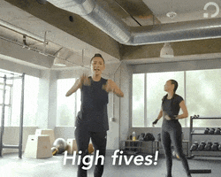 Video gif. Woman walks towards us and high fives the air with both her hands as she says, “High fives!”