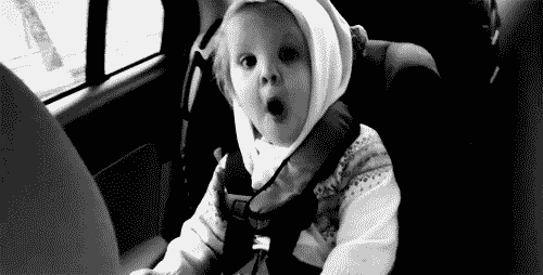 Video gif. Black and white footage of a toddler in a car seat with wide eyes and a gaping mouth as they shake excitedly.