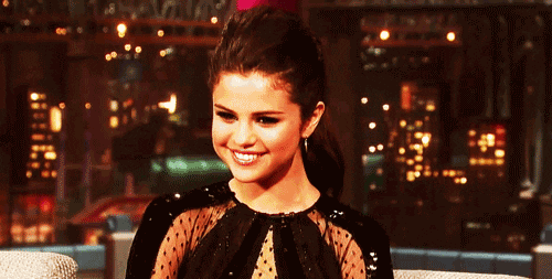 Celebrity gif. Selena Gomez smiles, facing downward as if flattered, at a talk show appearance.