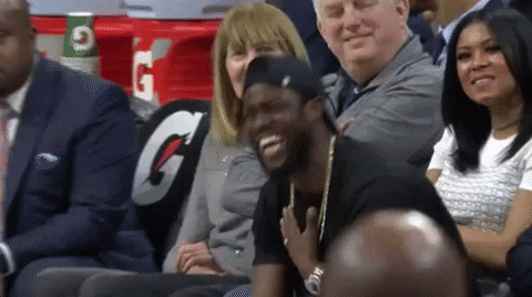 Celebrity gif. Kevin Hart sits on the sidelines at a basketball game holding a hand to his throat as he laughs uncontrollably. 