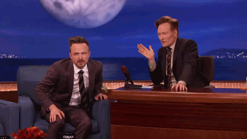 TV gif. Aaron Paul on Conan. He stands up and raises his hand in the air proudly.