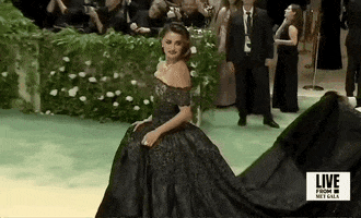 Met Gala 2024 gif. Penelope Cruz wearing a dark custom Chanel Couture off-shoulder gown, poses on the carpet, as someone fluffs her train into place.