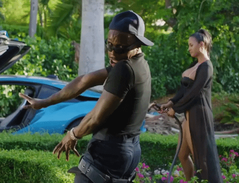 Music video gif. DaBaby poses in a garden with a blue sports car and a woman posing with a garden hose behind him.