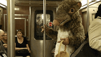 Video gif. A person dressed as a giant rat rides the subway, holding a basket.