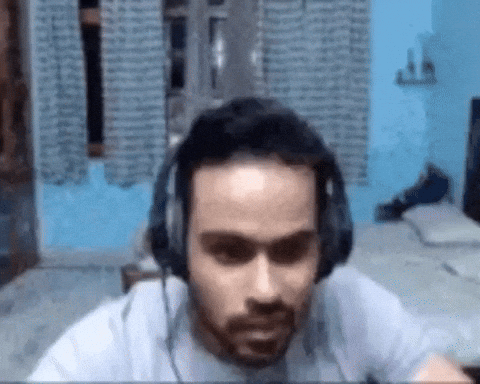 Video gif. A man wearing wired headphones and a gray t-shirt rolls back in his wheely chair, sticks out his tongue and wags his fingers in the air as he laughs to himself. 