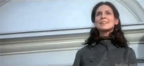 Movie gif. Holly Palance as a nanny in The Omen, standing on a ledge with a noose around her neck, looking honored.