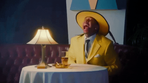 Music video gif. Tyga in Ayy Macarena wears a banana yellow suit and fedora at a table for one. He opens up his suit jacket to allow a heart made out of his white shirt to literally burst out into the open.