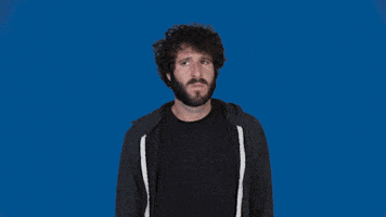 Celebrity gif. Lil Dicky stands in front of a solid blue background, looking from side to side with a slightly confused frown. He puts a hand to his chest as he speaks. Text, "Who me? He grins."