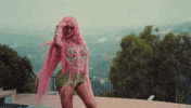 Music video gif. A scene from Saweetie's music video for "NANi" shows Saweetie on a hillside patio with long pink hair, a colorful beaded top and short lime green skirt surveying her surroundings with her hand over her eyes like a visor. 