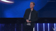 Video gif. Presenter Geoff Keighley smiles as he looks around at the audience from the 2024 Summer Game Fest stage. He is wearing a dark blue button-down shirt over a light gray t-shirt.