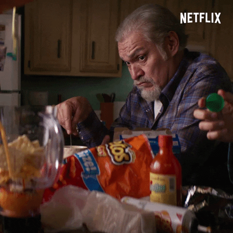 TV gif. Joaquín Cosío as Pop in Gentefied steeps a tea bag and looks with surprised but silent skepticism while watching someone fill a blender with various orange foods, on a counter littered with hot sauce, Cheetos, and crackers.