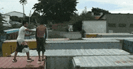 Video gif. Two boys stand on a shipping container. One boy jumps up and does a backflip over a gap between two shipping containers, and the other boy helps him make the jump by pushing him over mid-jump.