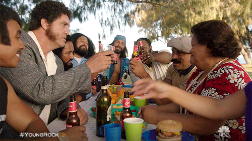 TV gif. A group of people, including Dwayne Johnson from Young Rock gather around a table holding bottles of beer. They smile and raise their bottles together, some of them saying, "cheers!"