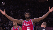 Sports gif. Joel Embiid of the Philadelphia 76ers walks across court, raising his arms to the side, scrunching his nose and nodding like he's feeling this moment.